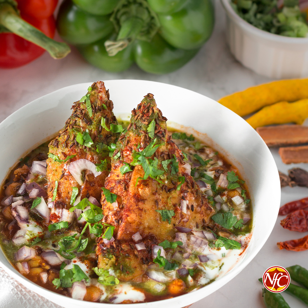 Samosa chaat in a white bowl: two crisp samosas over chickpeas with yogurt, tamarind and green chutney, red onion and fresh coriander