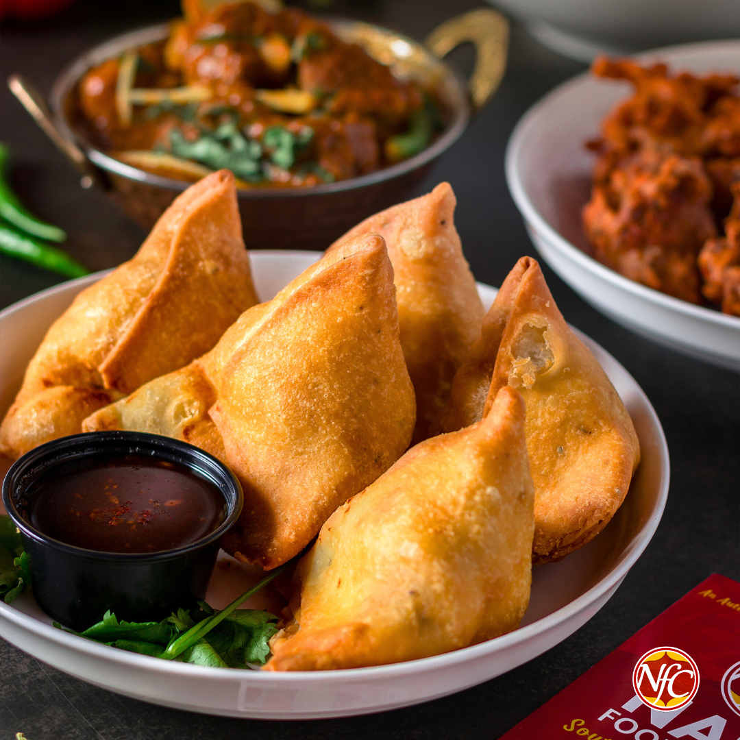 Bowl of vegetable samosas with tamarind chutney beside curry and pakoras