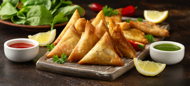 A wooden board of freshly fried samosas with red and green chutneys, lemon and chillies